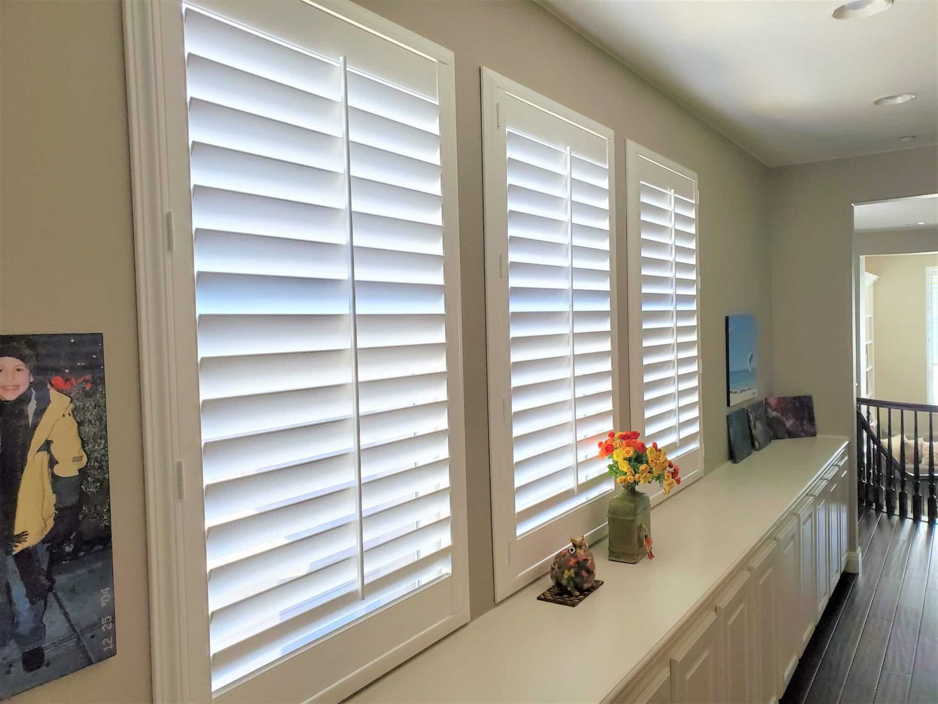 Three white shutters on windows in a hallway, with flowers and decorations on a white counter.