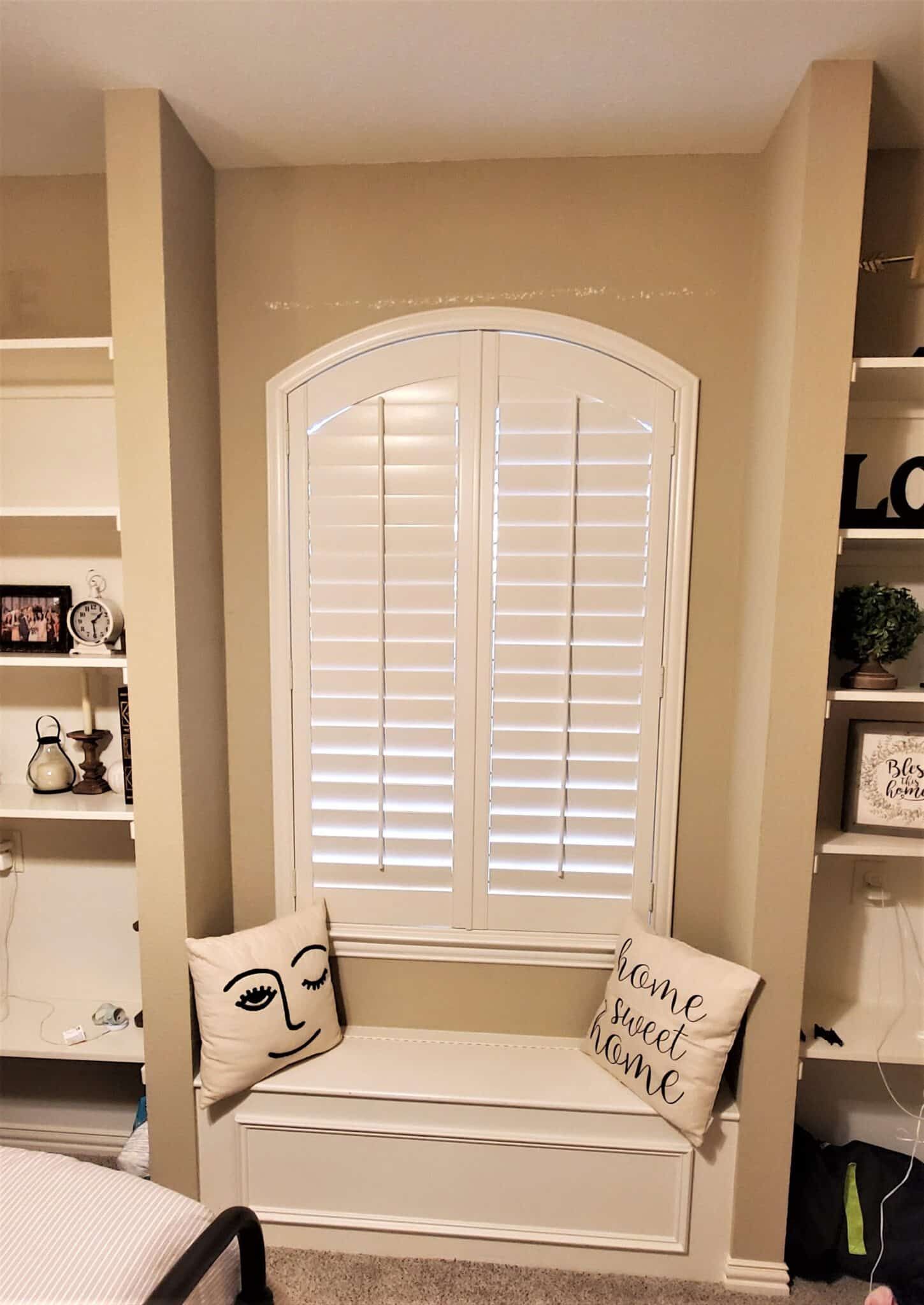 Window seat with white shutters, built-in shelves, tan walls, and two decorative pillows.