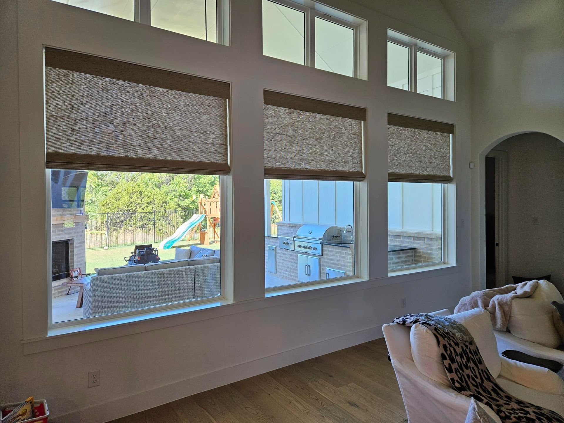 Living room with three large windows, each with a woven shade, overlooking a backyard with a playground.
