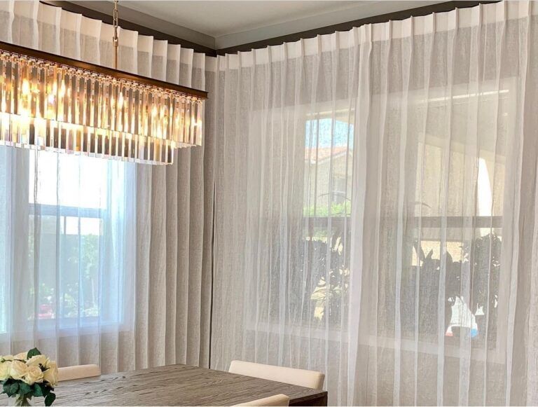 Sheer white curtains frame windows in a well-lit dining room, with a chandelier and table set for a meal.