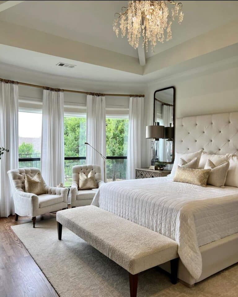 Elegant bedroom with neutral tones; white tufted headboard, crystal chandelier, and large windows.