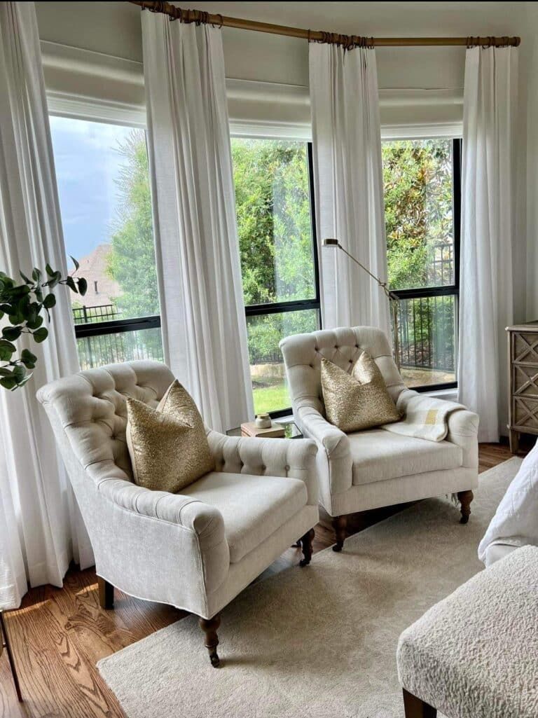 Two white tufted armchairs with gold pillows, facing windows with white curtains, in a well-lit room.