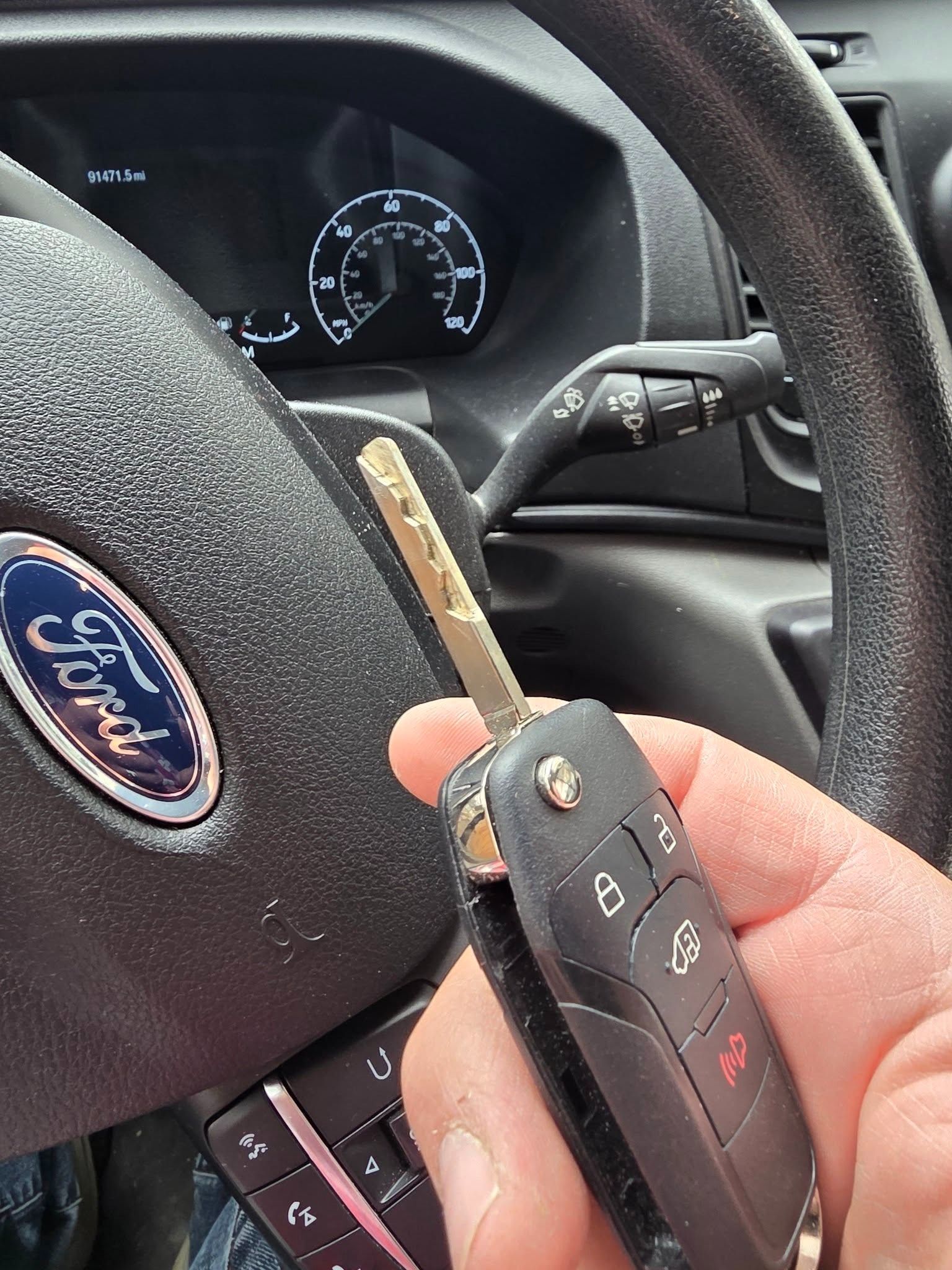 Hand holding a Ford car key fob beside a Ford steering wheel and dashboard gauges