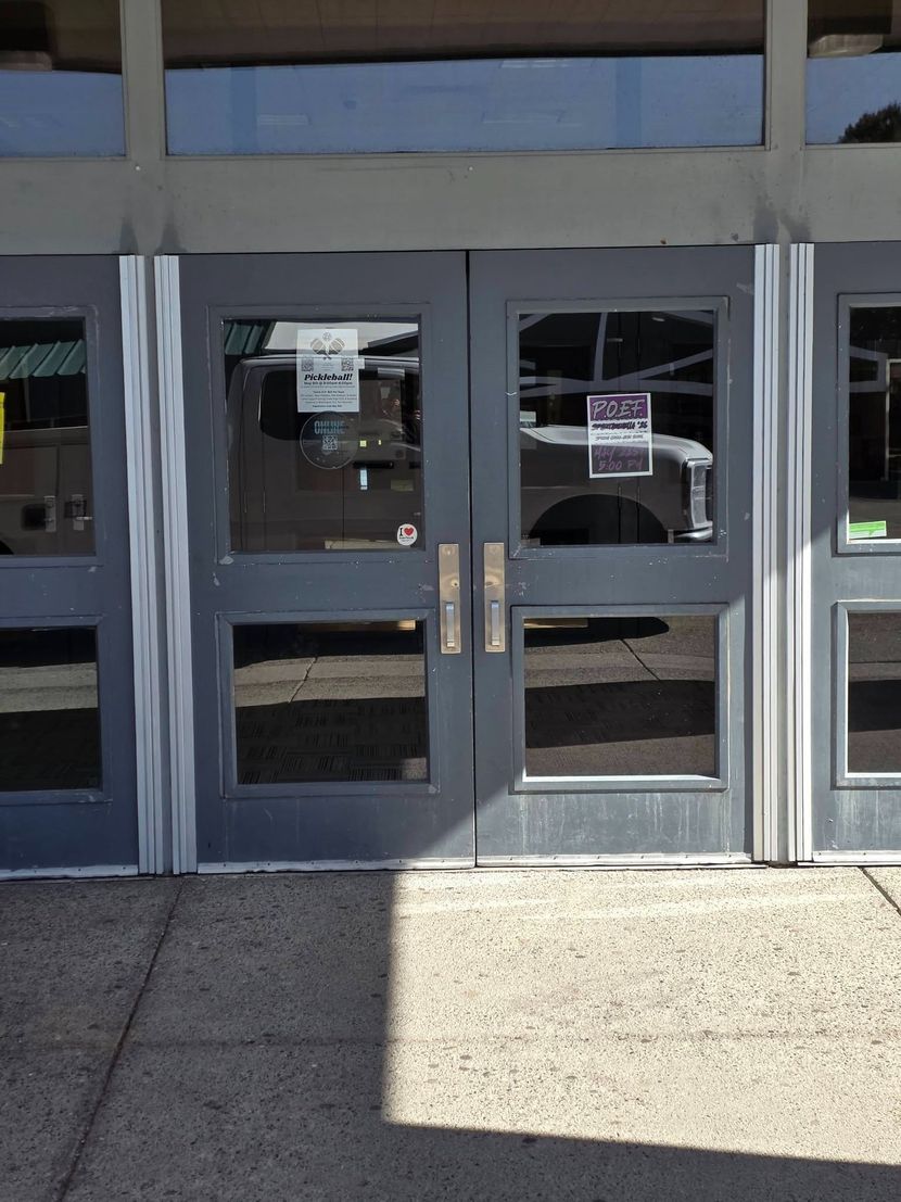Glass double doors at a building entrance with posted notices and sunlight on the sidewalk