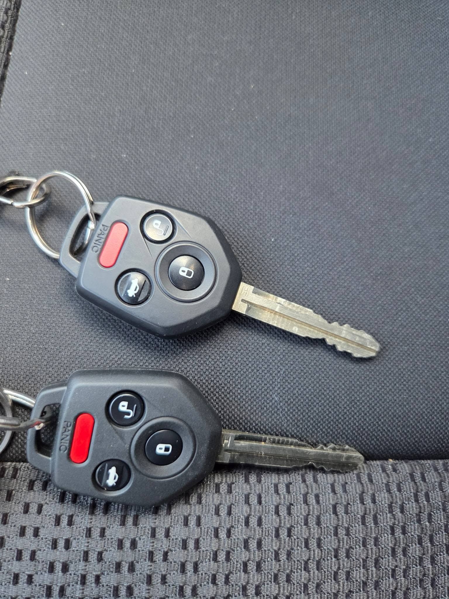 Two car key fobs on a gray textured fabric surface, one black with a red button and one silver trim.