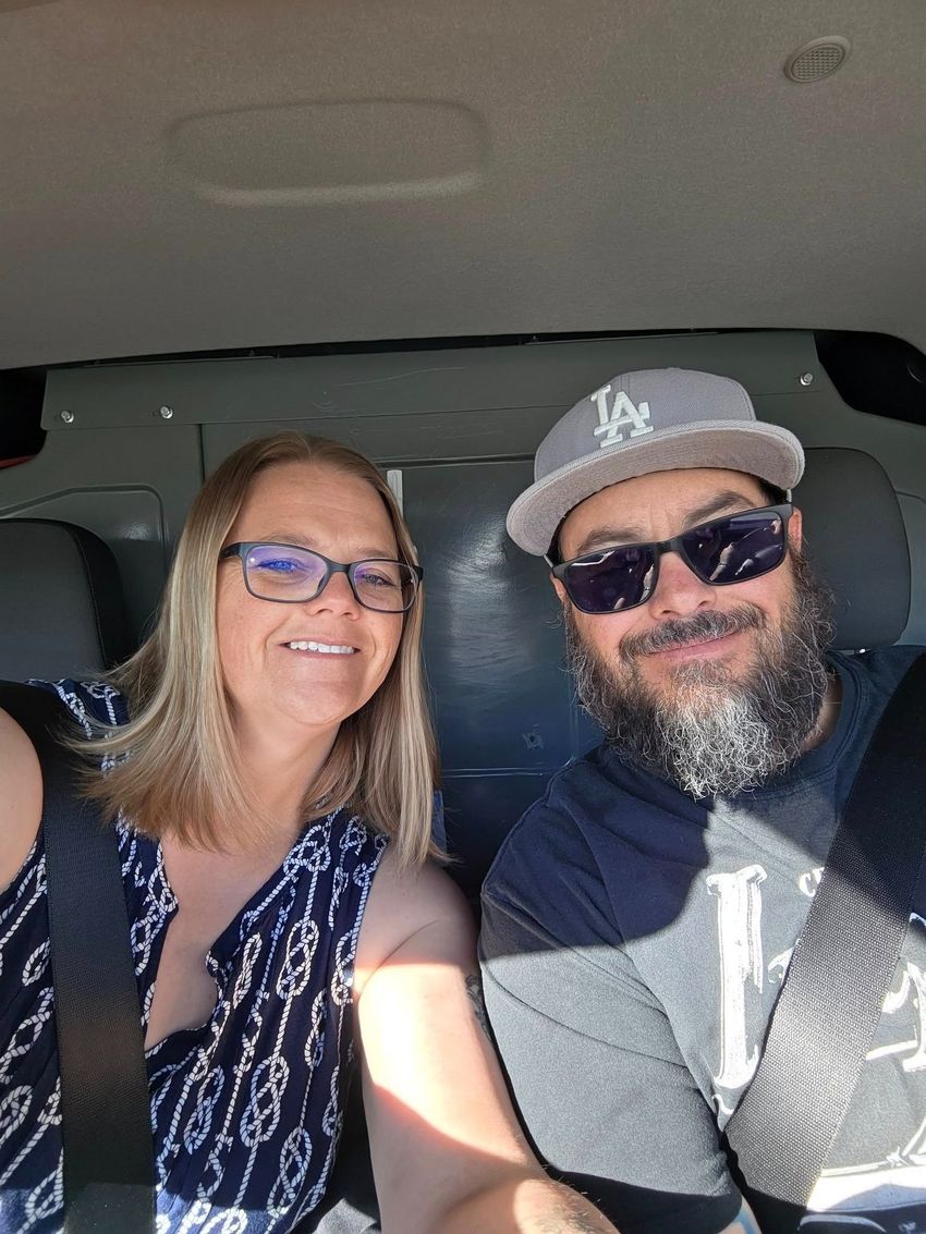 Two smiling people in a car, wearing sunglasses and casual clothes.