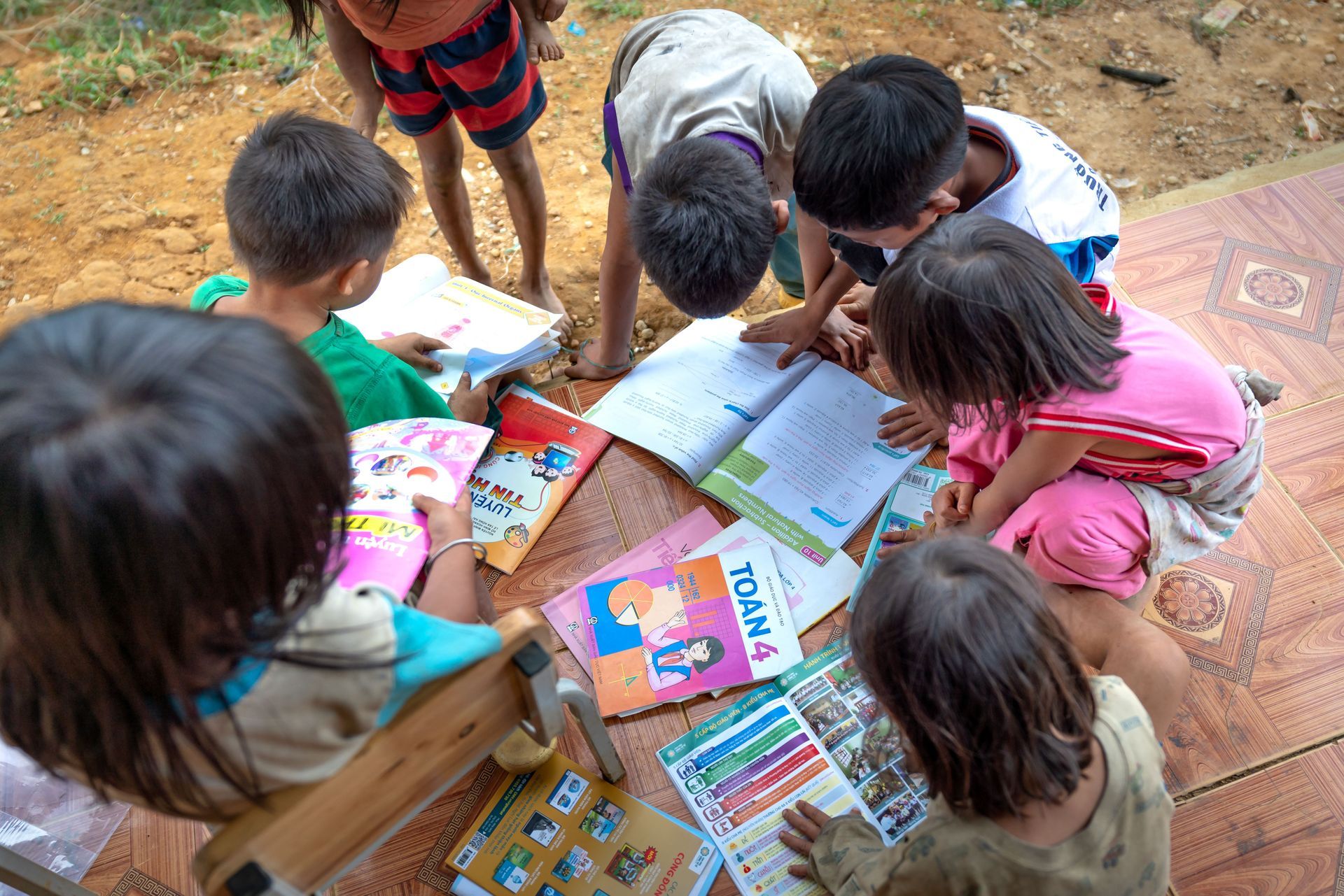Children gathered outdoors around books and notebooks, reading and studying together on the ground.