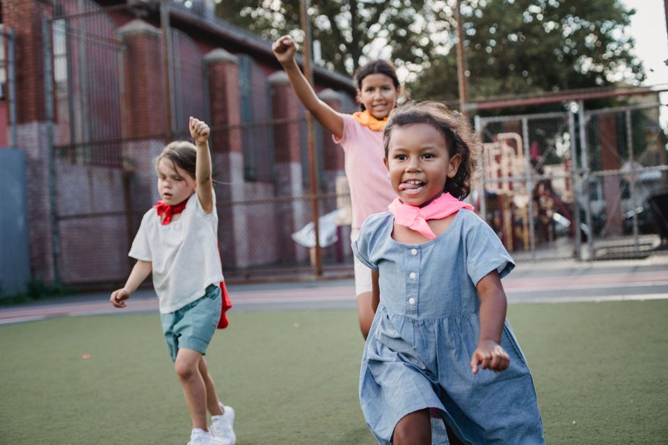 Children running and smiling on a playground, with one girl in the foreground in a blue dress