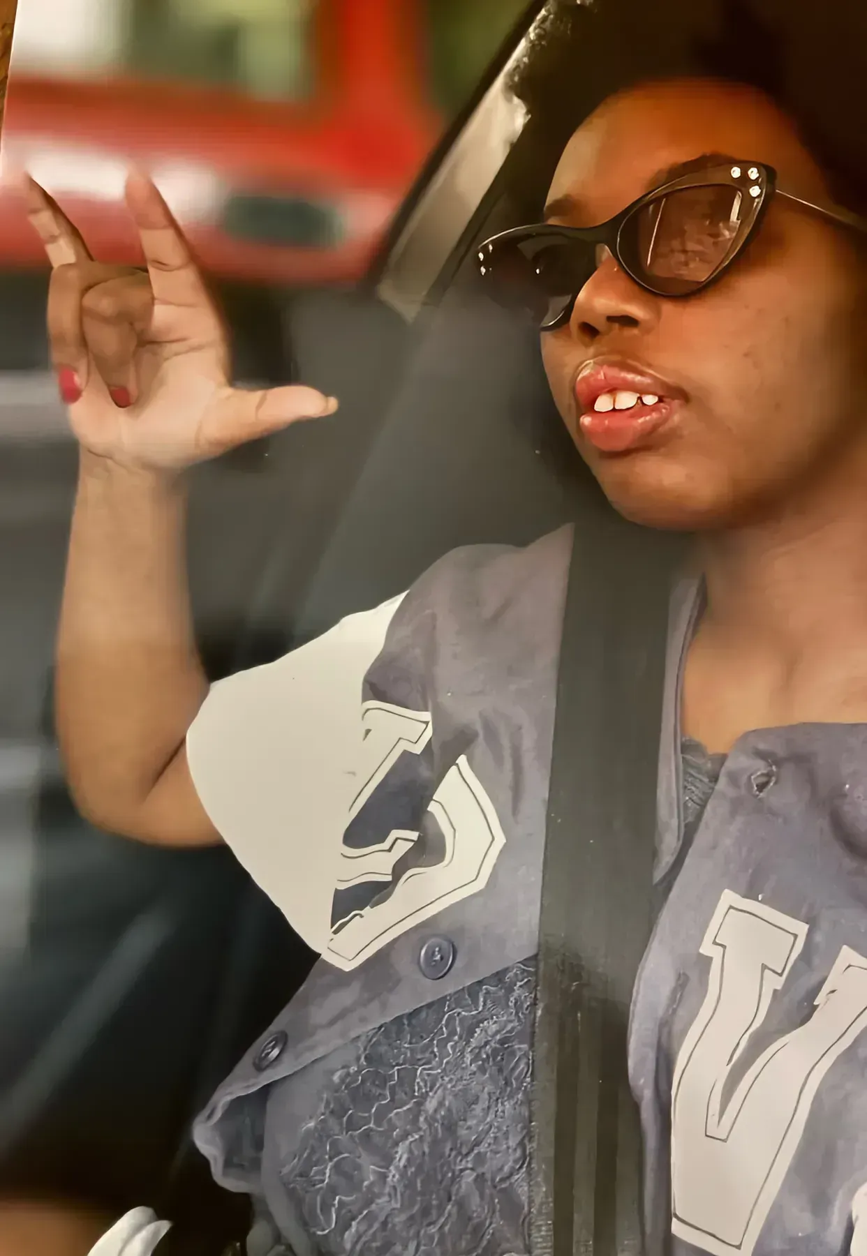 Person wearing sunglasses and a seatbelt makes a hand gesture inside a car.