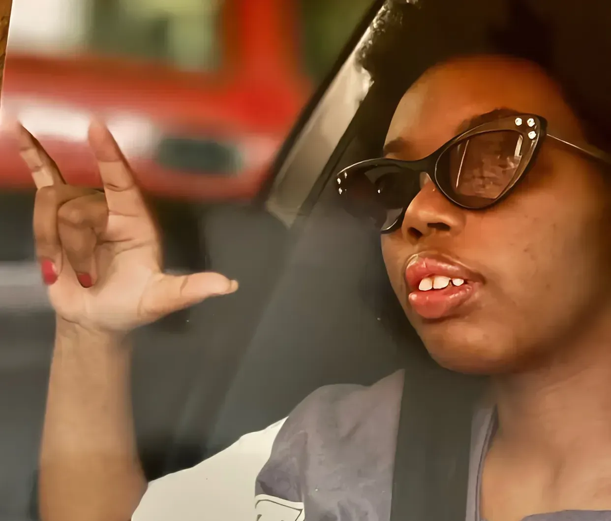 Person wearing sunglasses and a seat belt, making a hand gesture inside a car.