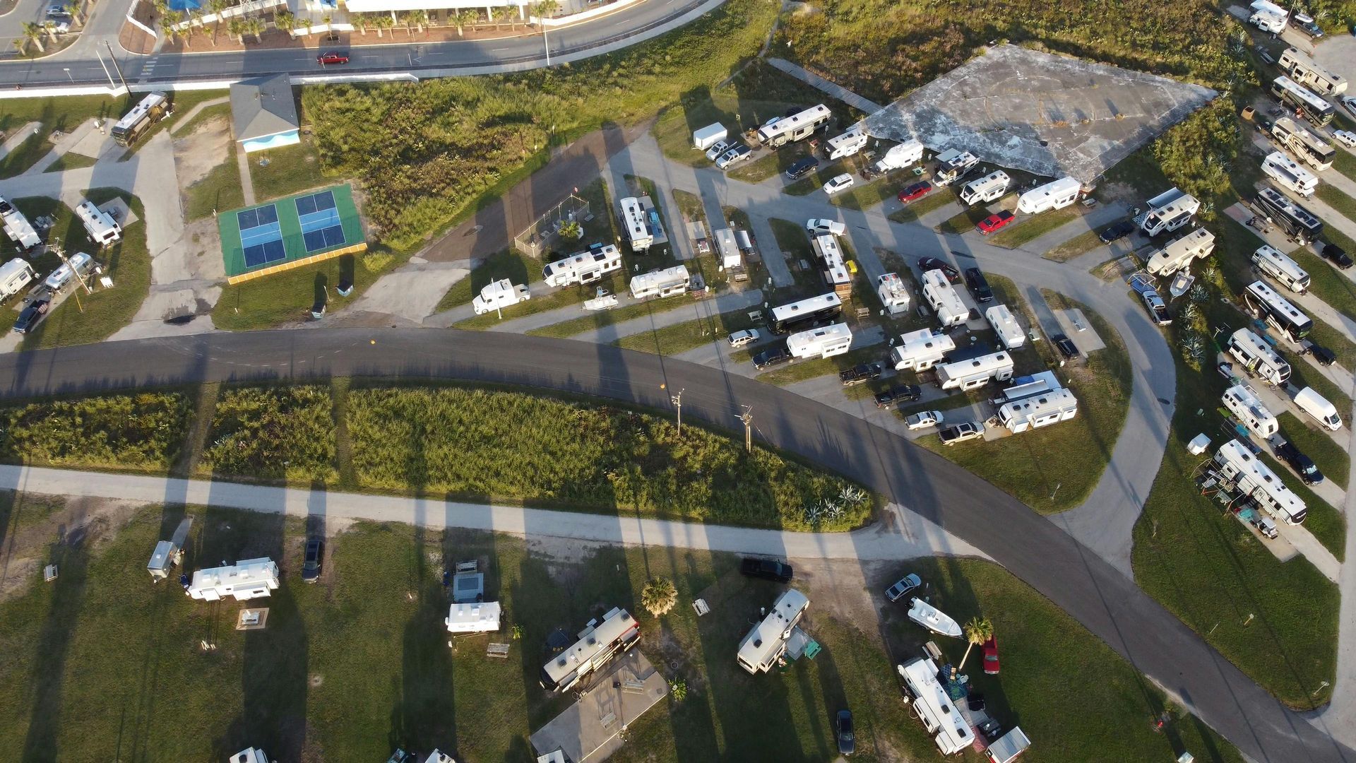 Aerial view of a parking lot, curving roads, and tennis courts beside a wooded area.
