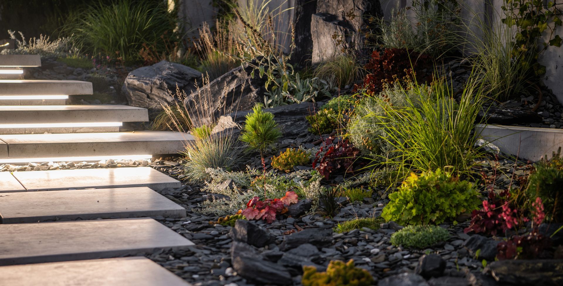 Stone walkway beside a landscaped garden with rocks, shrubs, and colorful flowers in sunlight