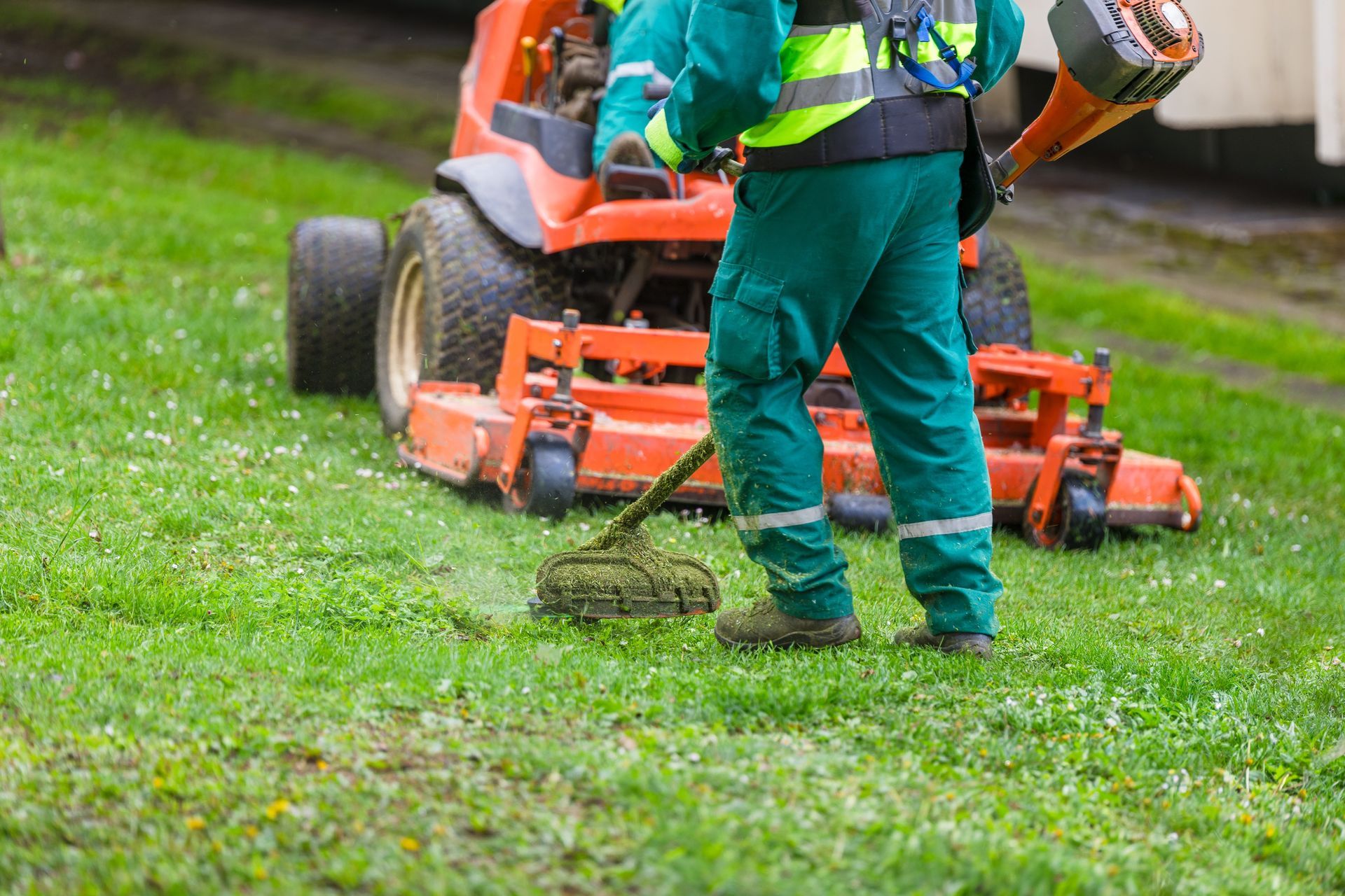 Person mowing a green lawn with an orange ride-on mower while wearing teal work clothes.