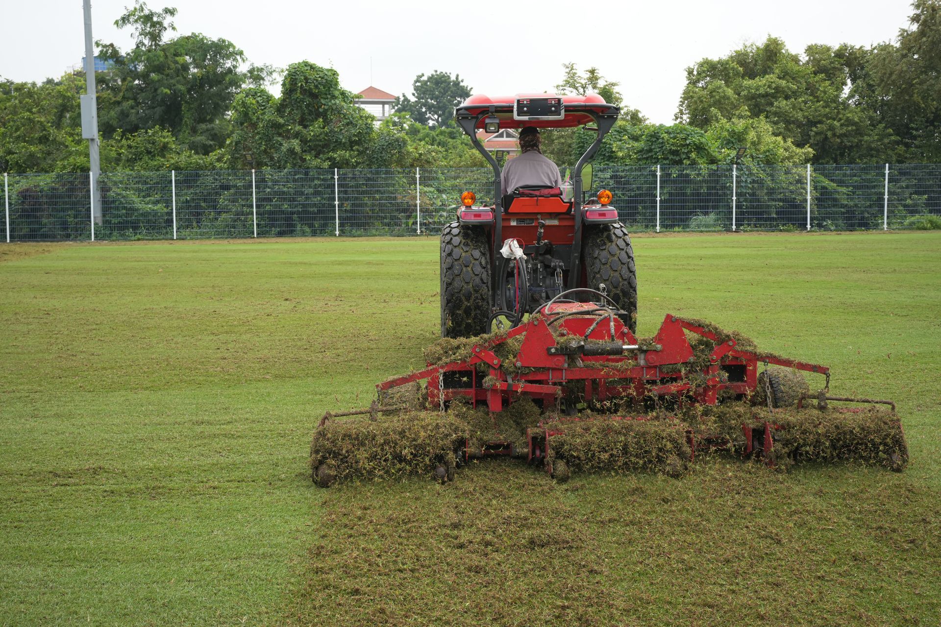 Tractor mowing a green field with a red rotary cutter near a fence and trees
