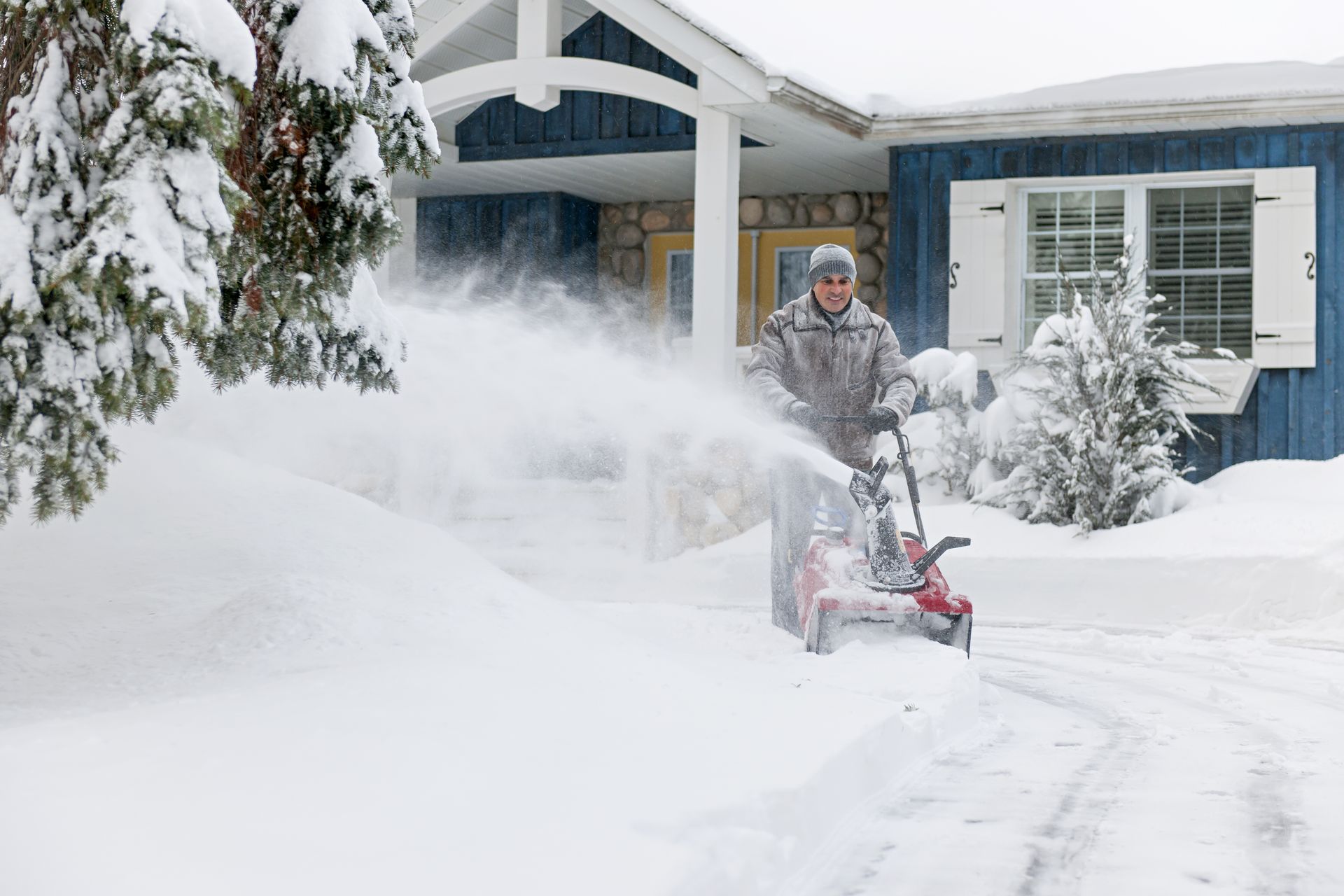Person using a snowblower clearing a snowy driveway in front of a house.