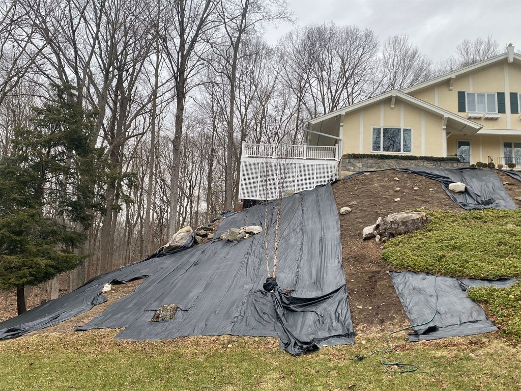 House on a steep hillside with black erosion-control tarp and bare trees in the background