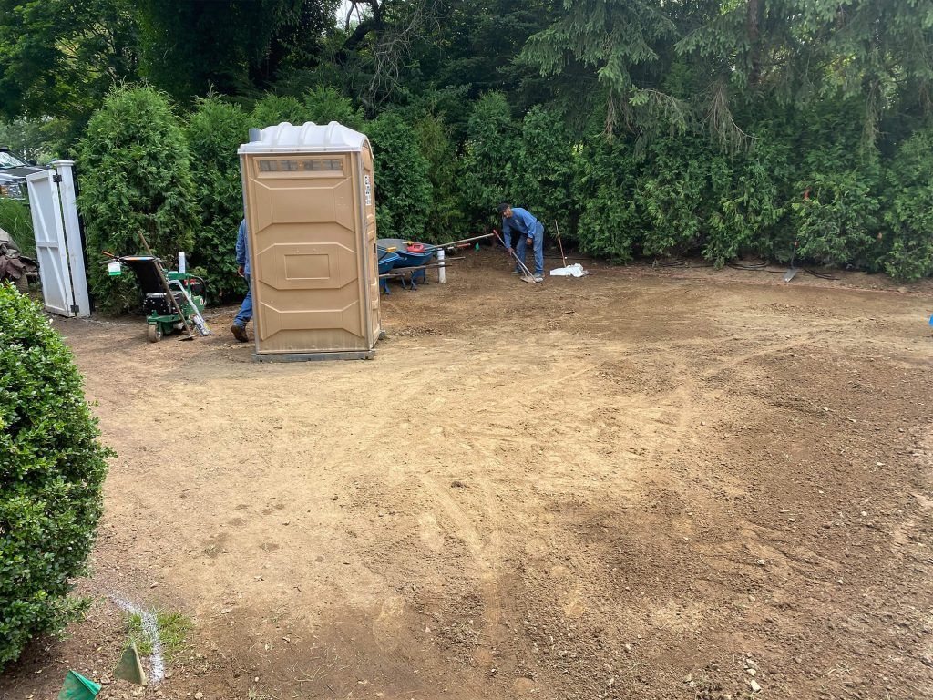 Outdoor dirt lot with a portable toilet, trees, and a person working near the back.