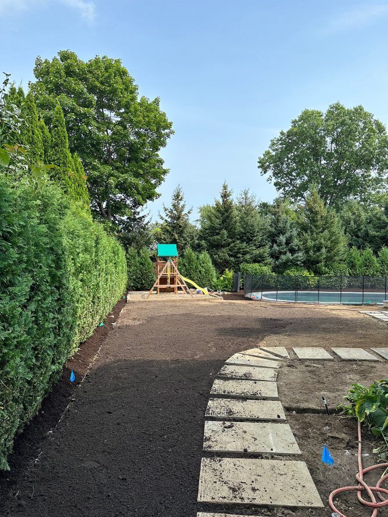 Backyard playground with a wooden playset, mulch ground, stone path, and tall green trees under a blue sky
