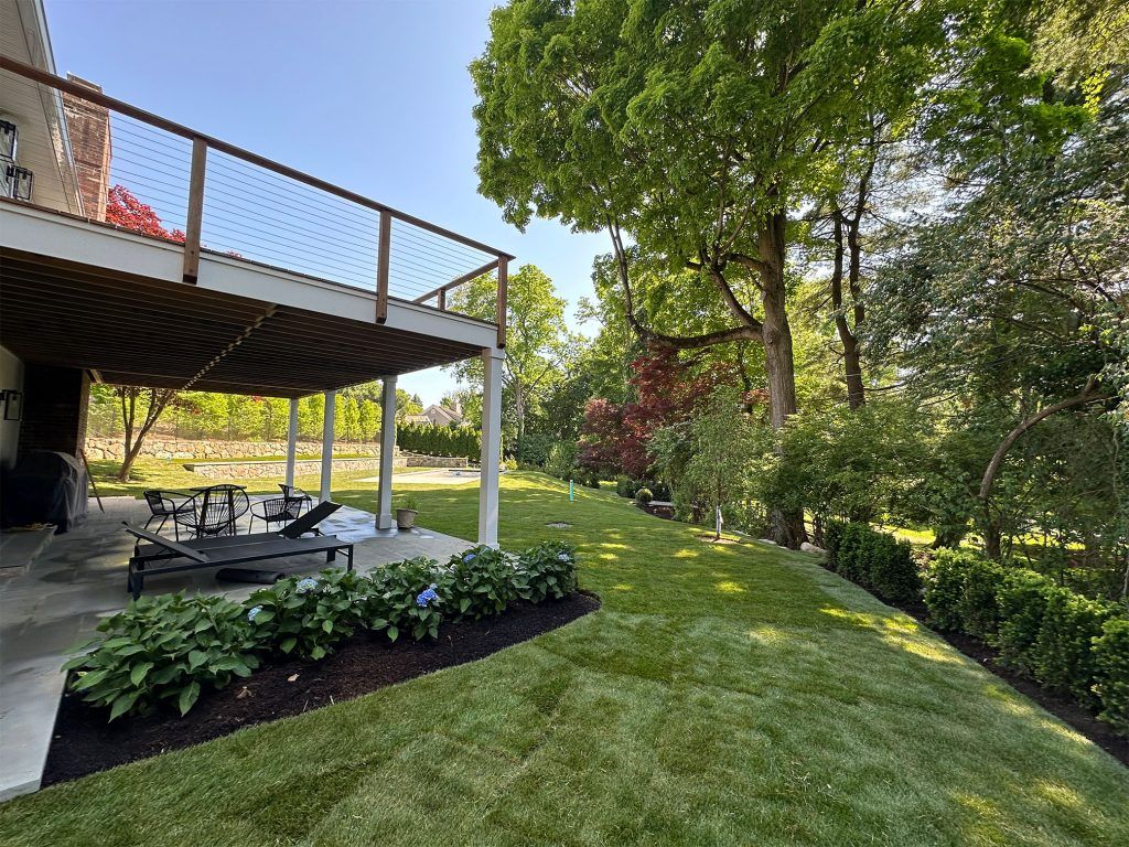 Backyard deck with stairs, patio furniture, and a green lawn bordered by trees and shrubs.