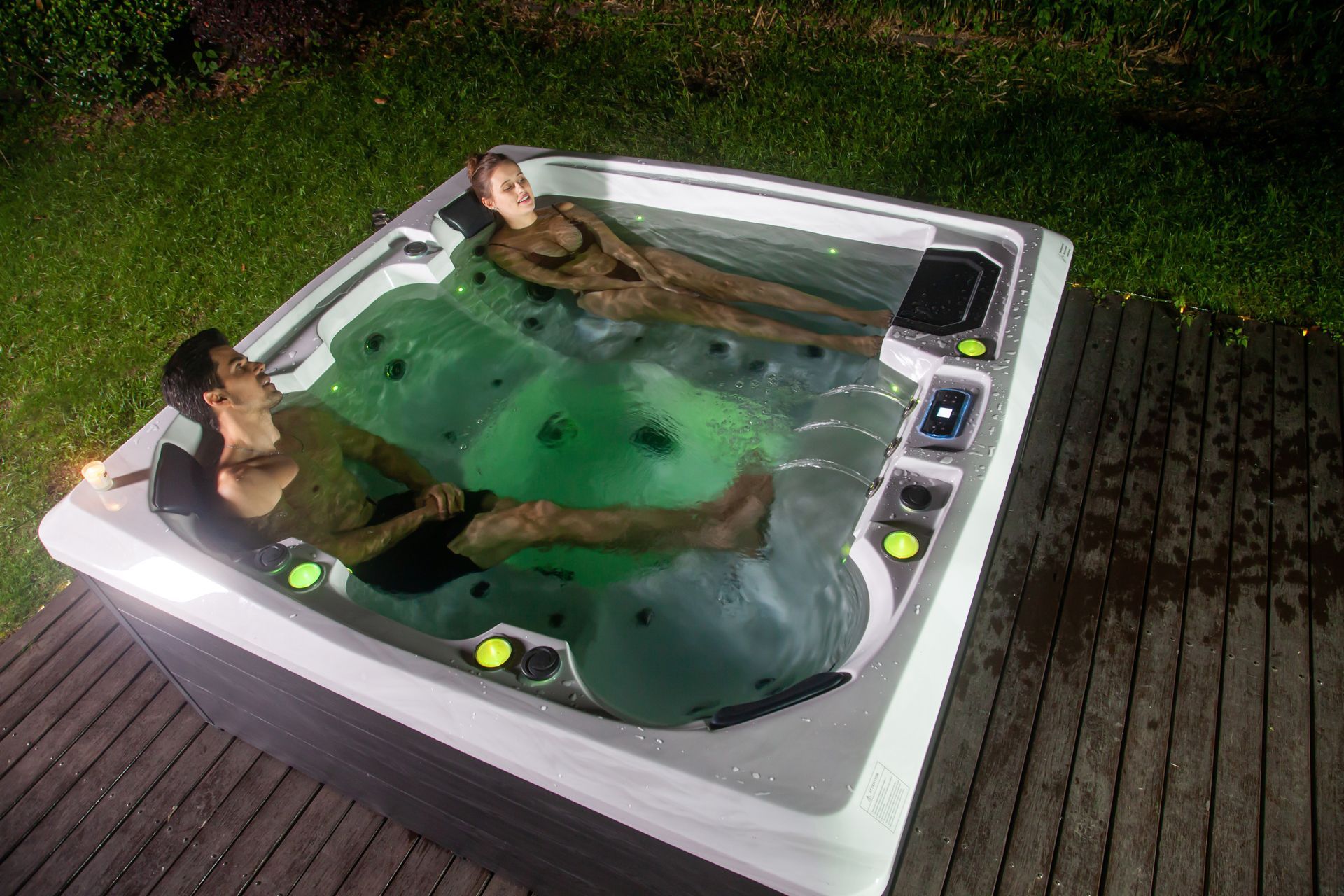 Two people relaxing in a lit hot tub on a wooden deck at night