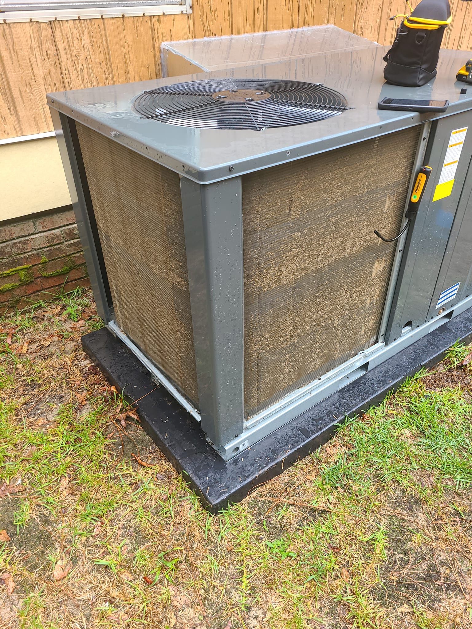 HVAC unit outside a building, with visible fan and dirty coil filter, sitting on a black base.