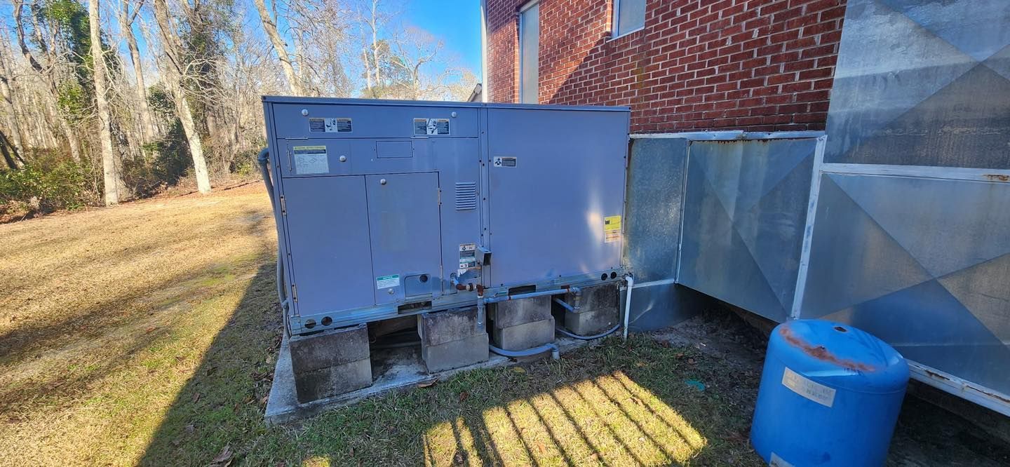 An exterior air conditioning unit next to a brick building. Blue plastic drum in the foreground, dead grass.