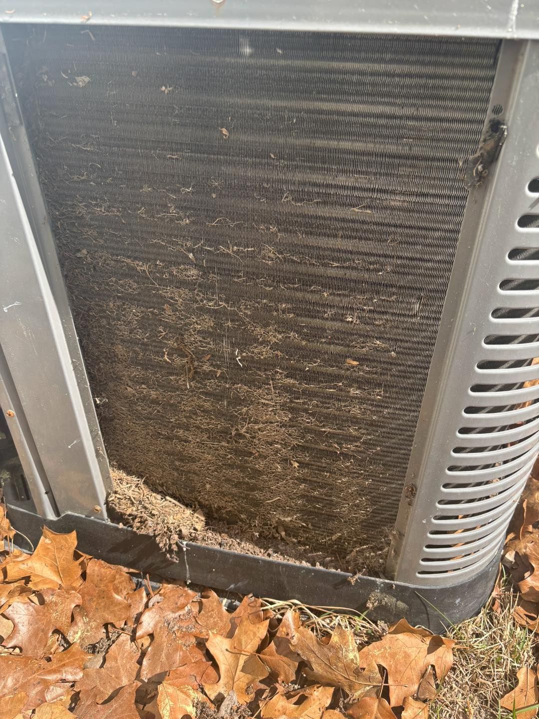 Dirty air conditioner coil packed with debris. Grey metal unit surrounded by fallen leaves.