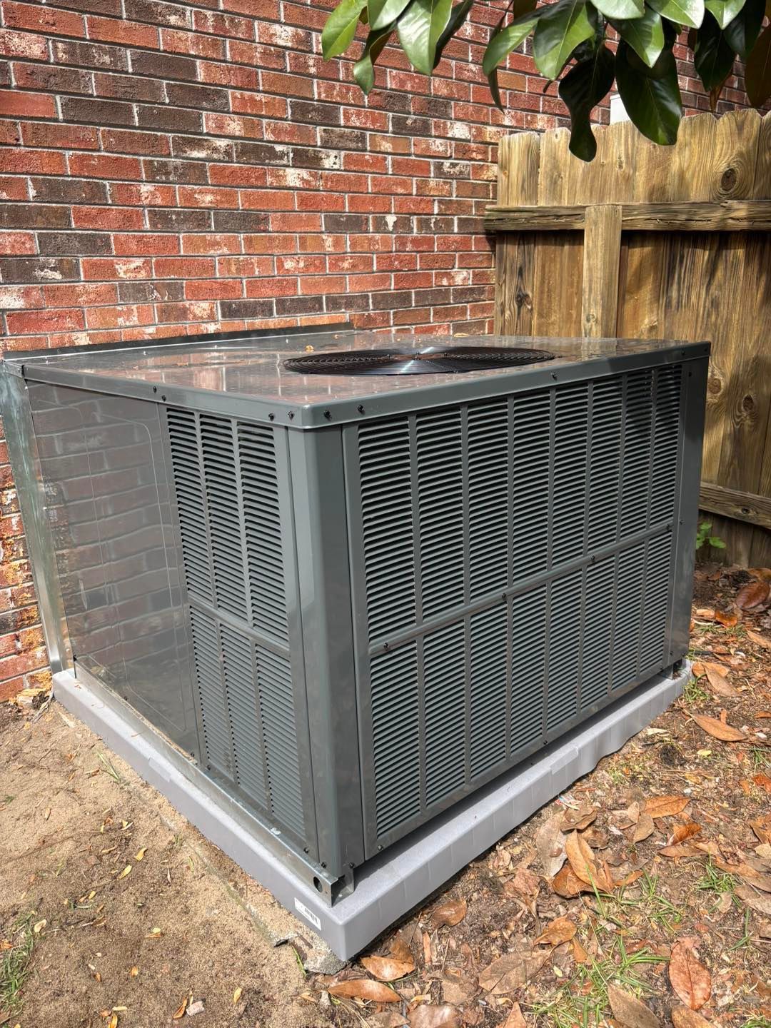 Gray air conditioning unit outside, against a brick wall and wooden fence.