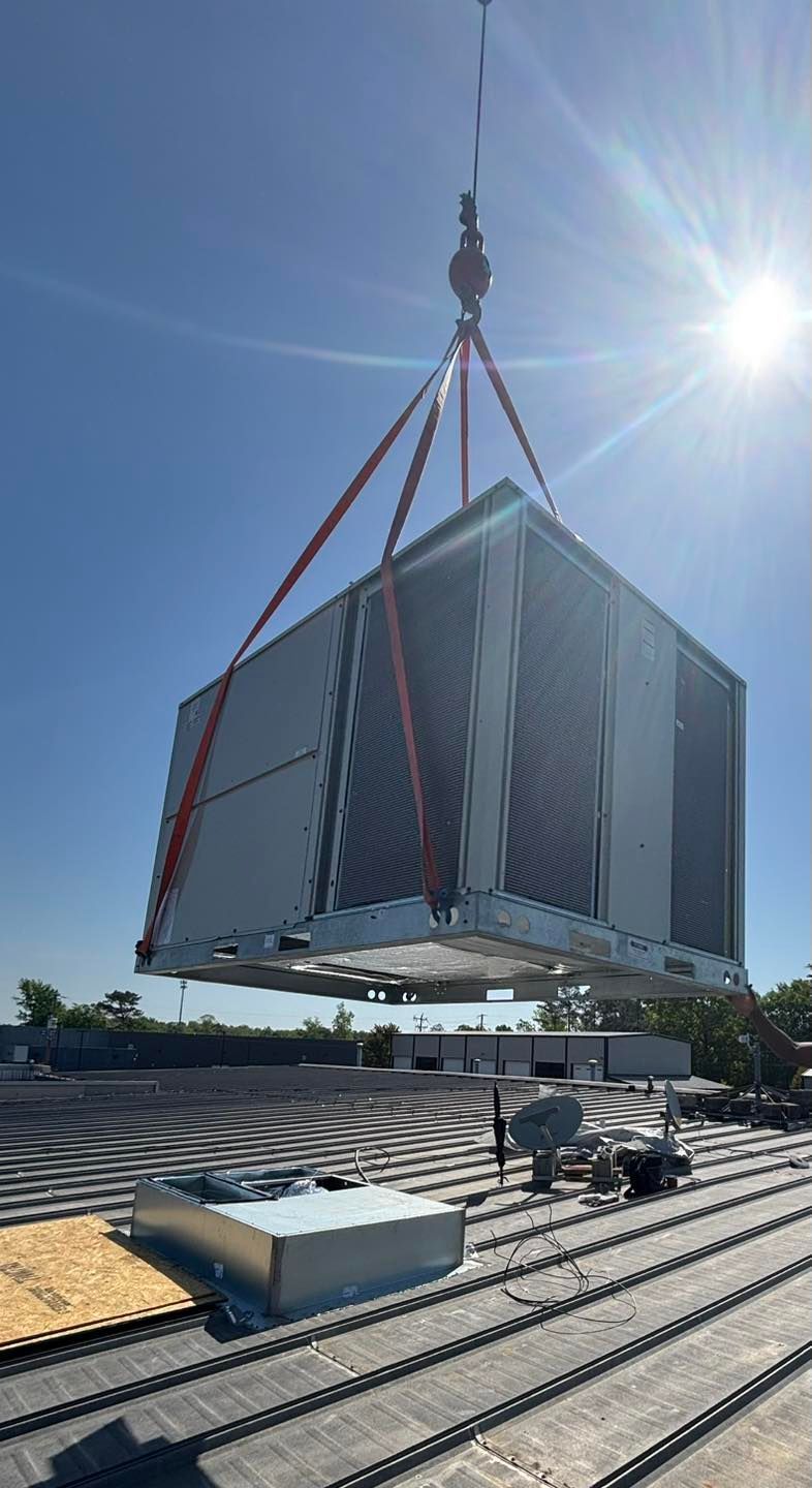 A large HVAC unit being lifted by a crane onto a building rooftop on a sunny day.