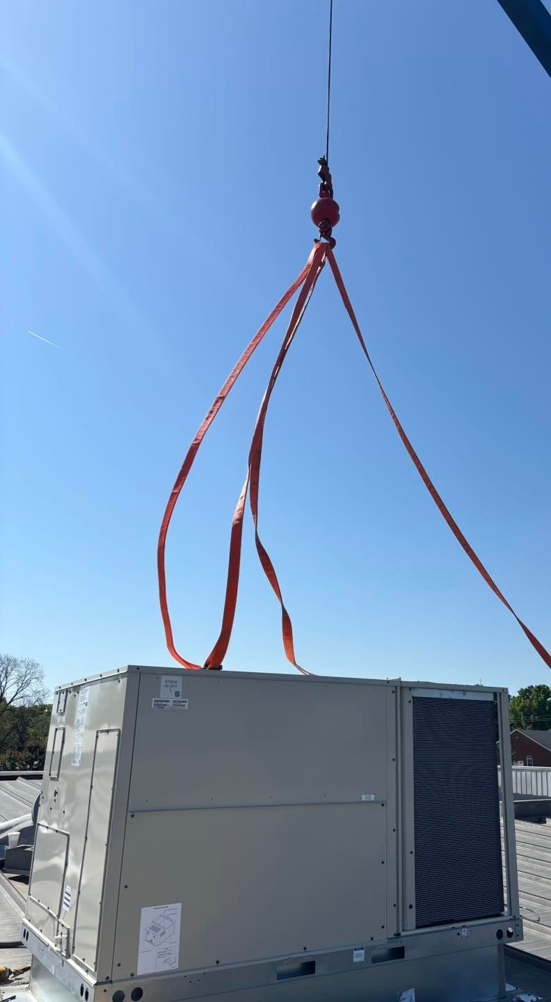A rooftop HVAC unit being lifted by red straps from a crane against a blue sky.