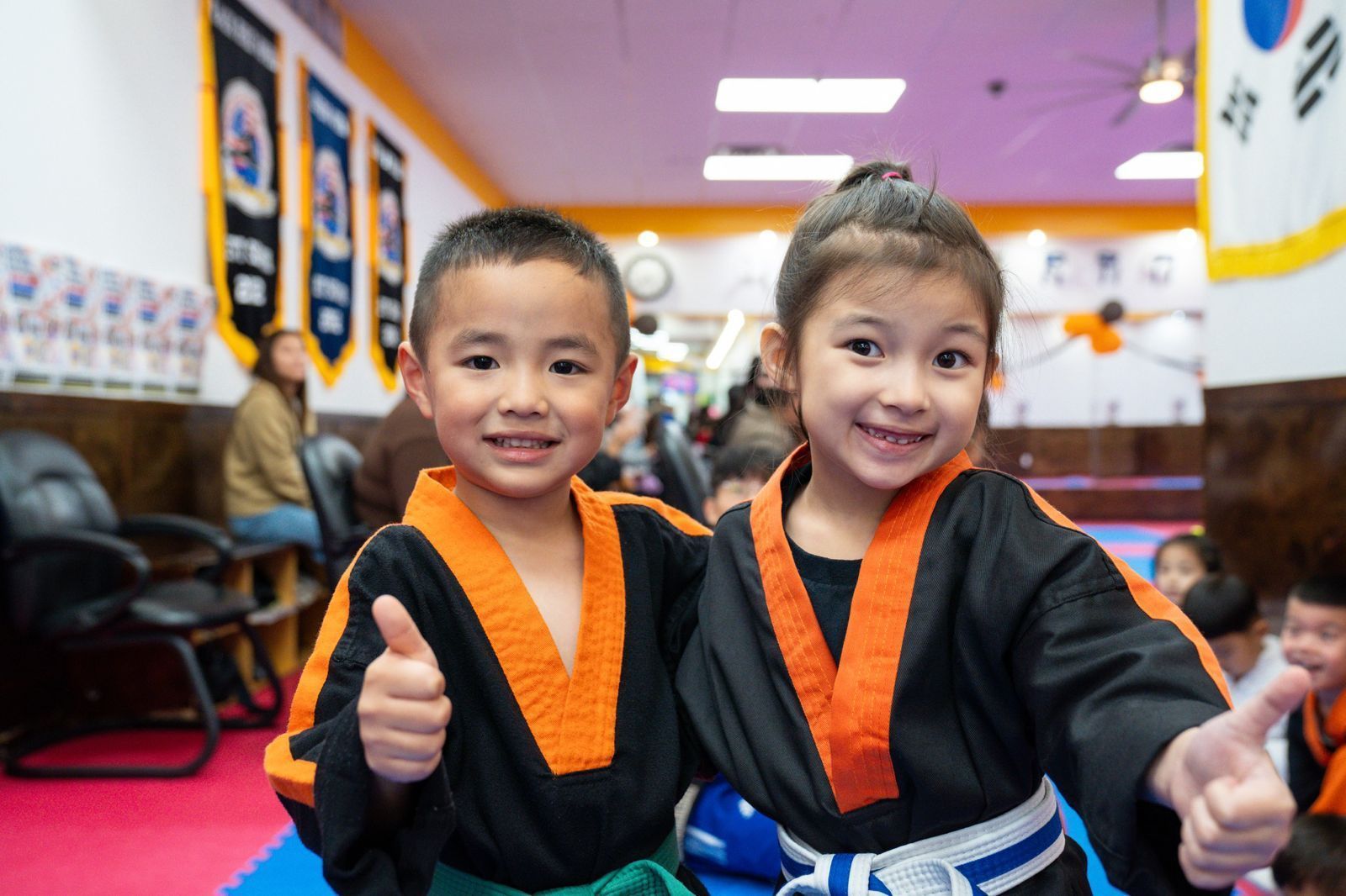 A boy and a girl in karate uniforms are giving a thumbs up in a gym.