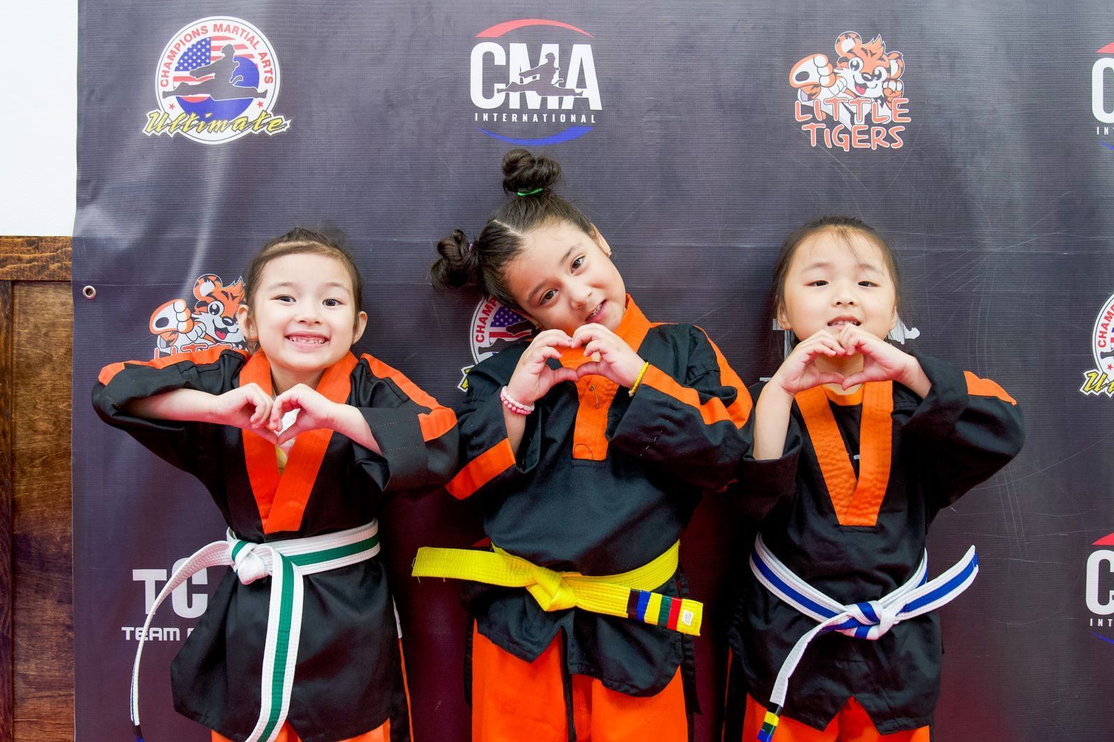 Three young girls in martial arts uniforms are making hearts with their hands.