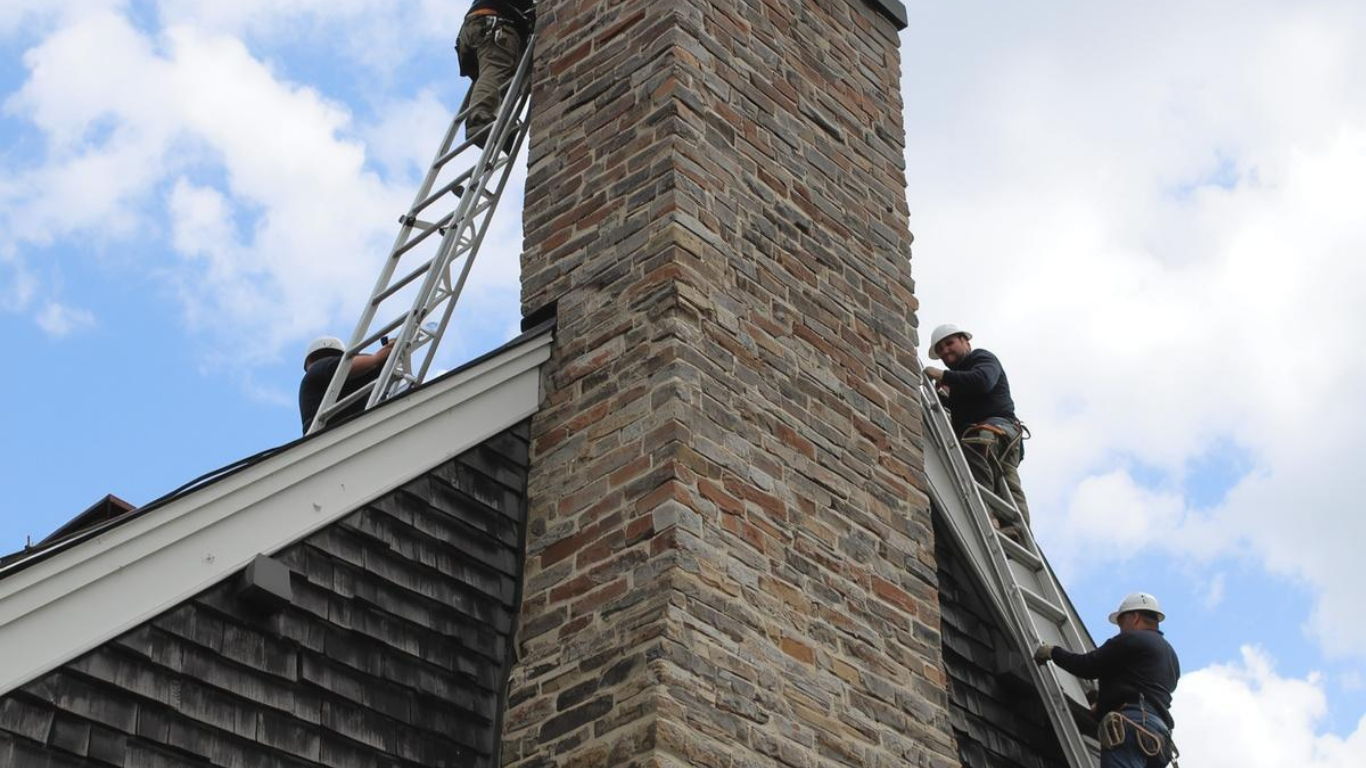 Three workers in hard hats and safety harnesses use ladders to work on a tall stone chimney on a shingled roof.