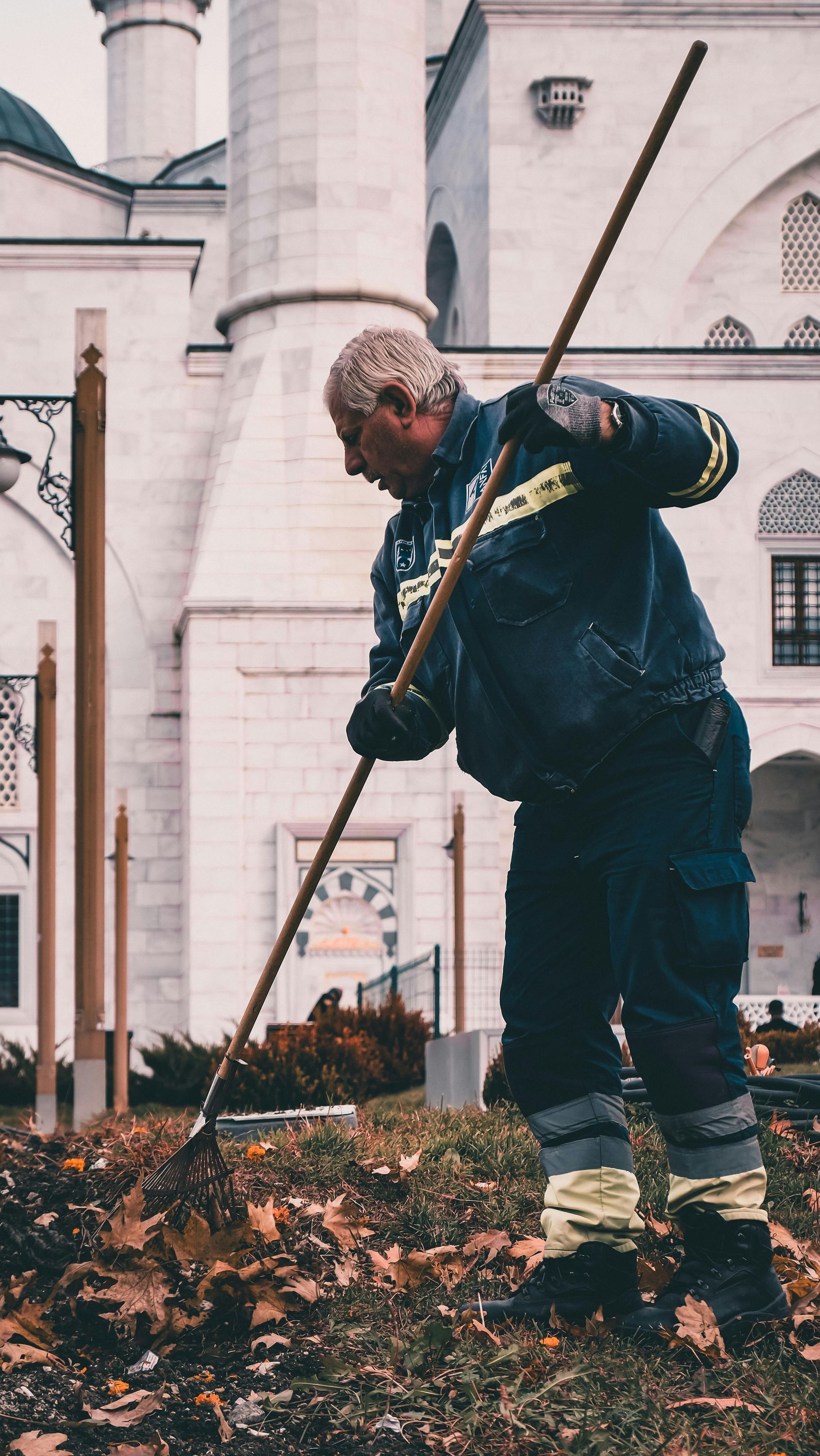 Man raking leaves near a mosque. He's wearing blue work clothes and high-visibility boots.