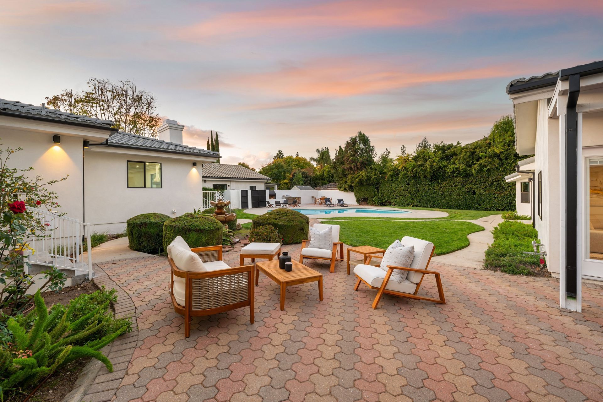Patio with wooden furniture, brick pavers, and a view of a lawn and pool, under a sunset sky.