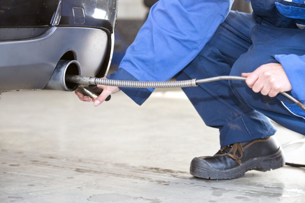 Emissions Repair and Diagnosis Mechanic in blue overalls testing car emissions with a hose in a garage.