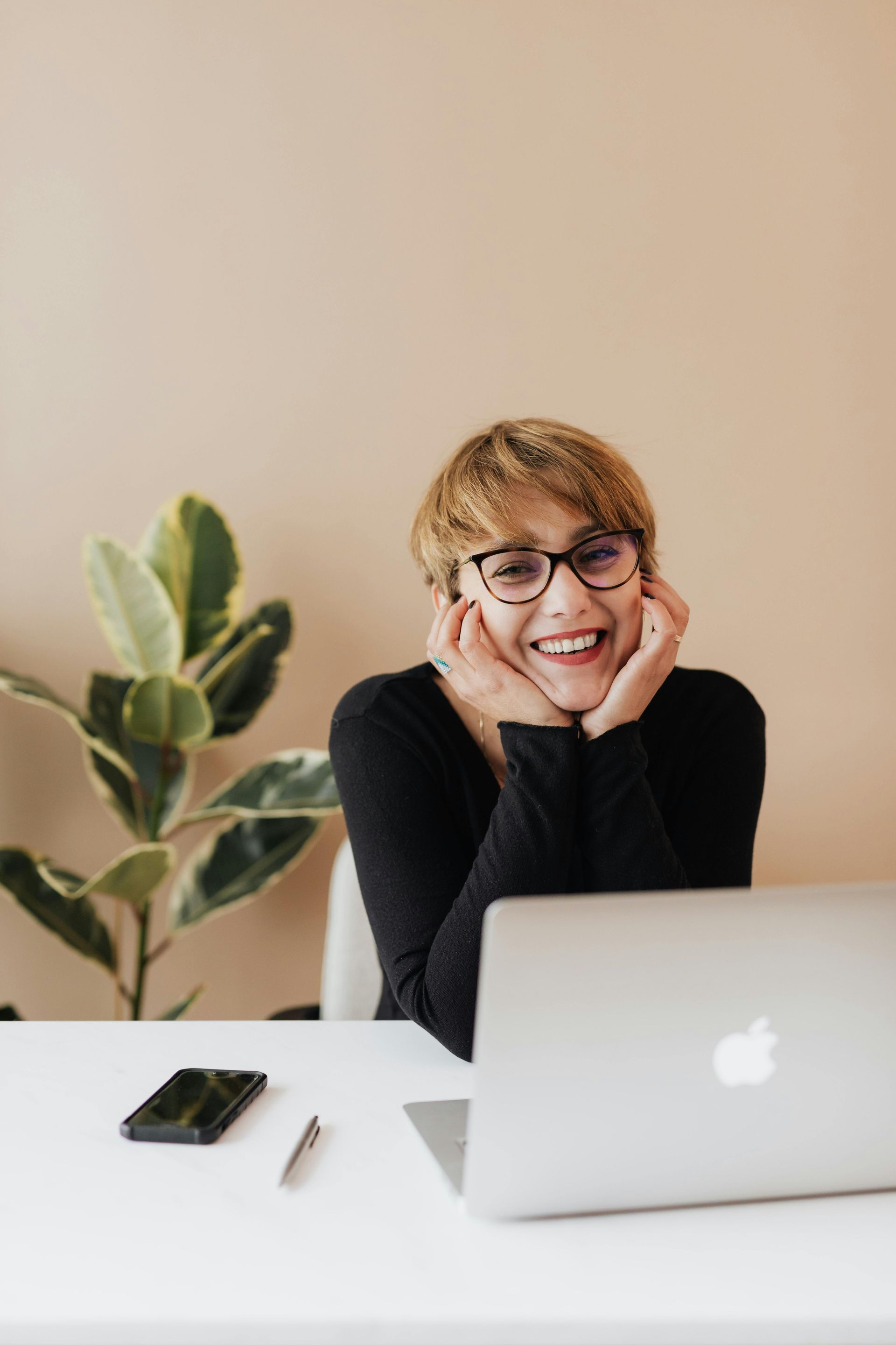 Person smiling at a desk with a laptop, phone, and plant against a beige wall.