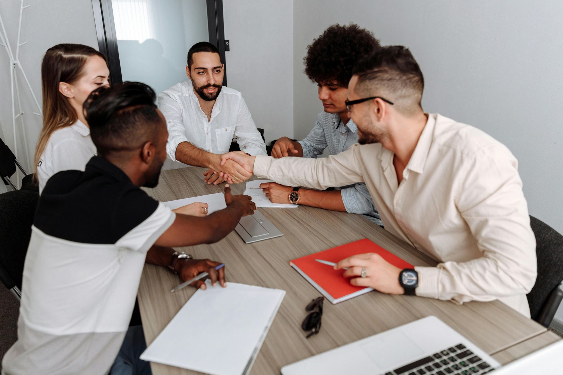 Team meeting around a conference table, with colleagues smiling and shaking hands over laptops and notebooks