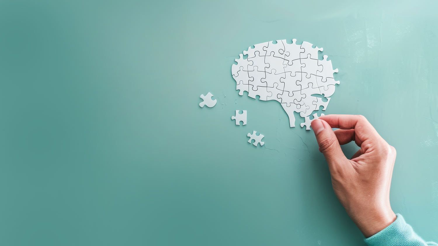 Hand fitting puzzle pieces to a brain shape on a teal background.