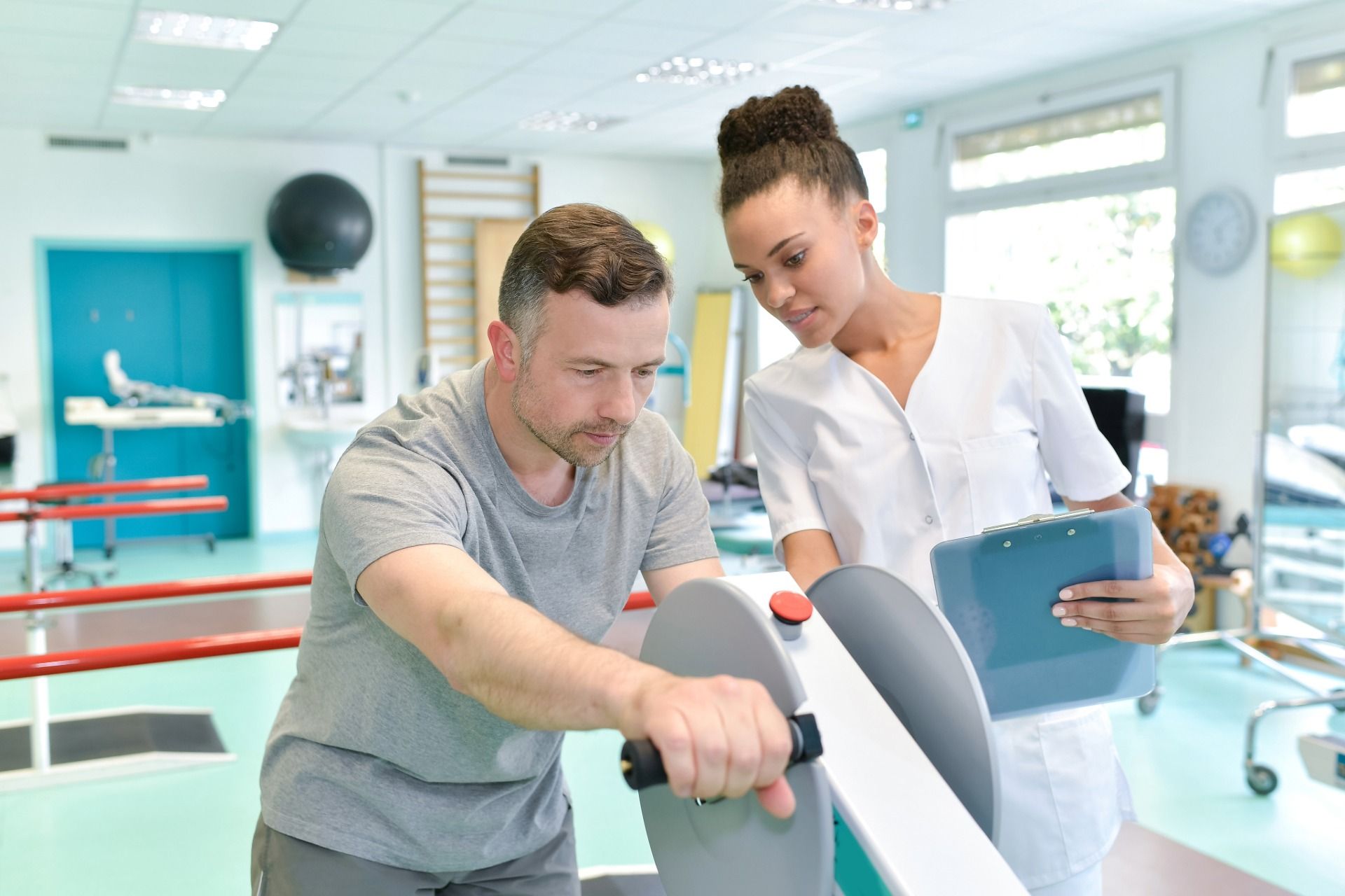 Man using exercise equipment, guided by a healthcare professional in a rehab setting.