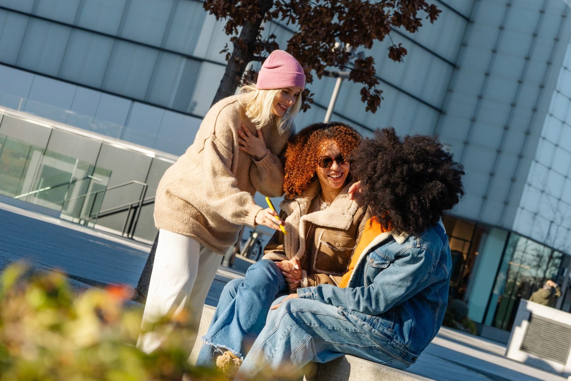 Three people sit and stand outside, talking. One wears a pink hat, another a brown jacket, and one a denim jacket.