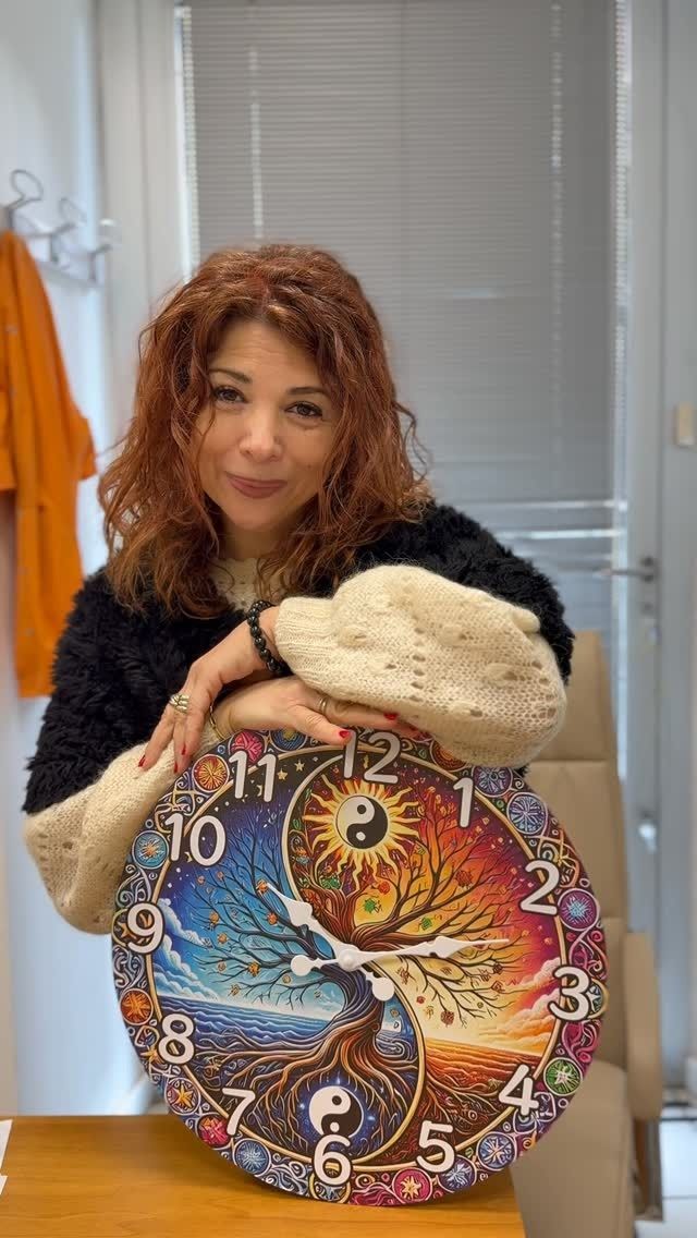 Una donna tiene in mano un dipinto circolare dai colori vivaci, con motivi celestiali e di orologi, in un ambiente interno vicino a una finestra.