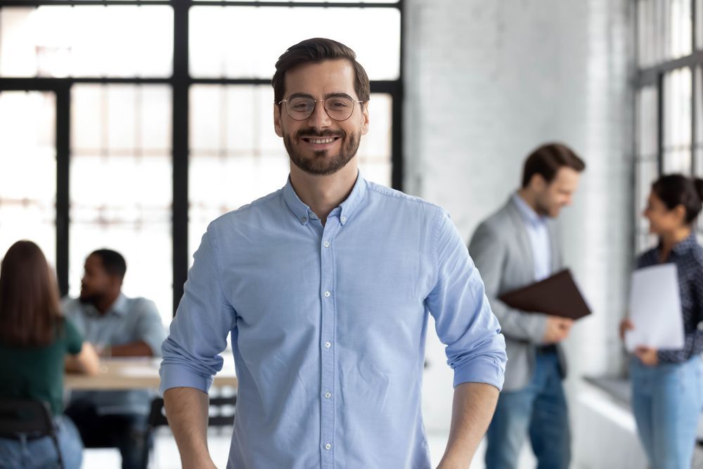 A man in a blue shirt and glasses is standing while smiling.