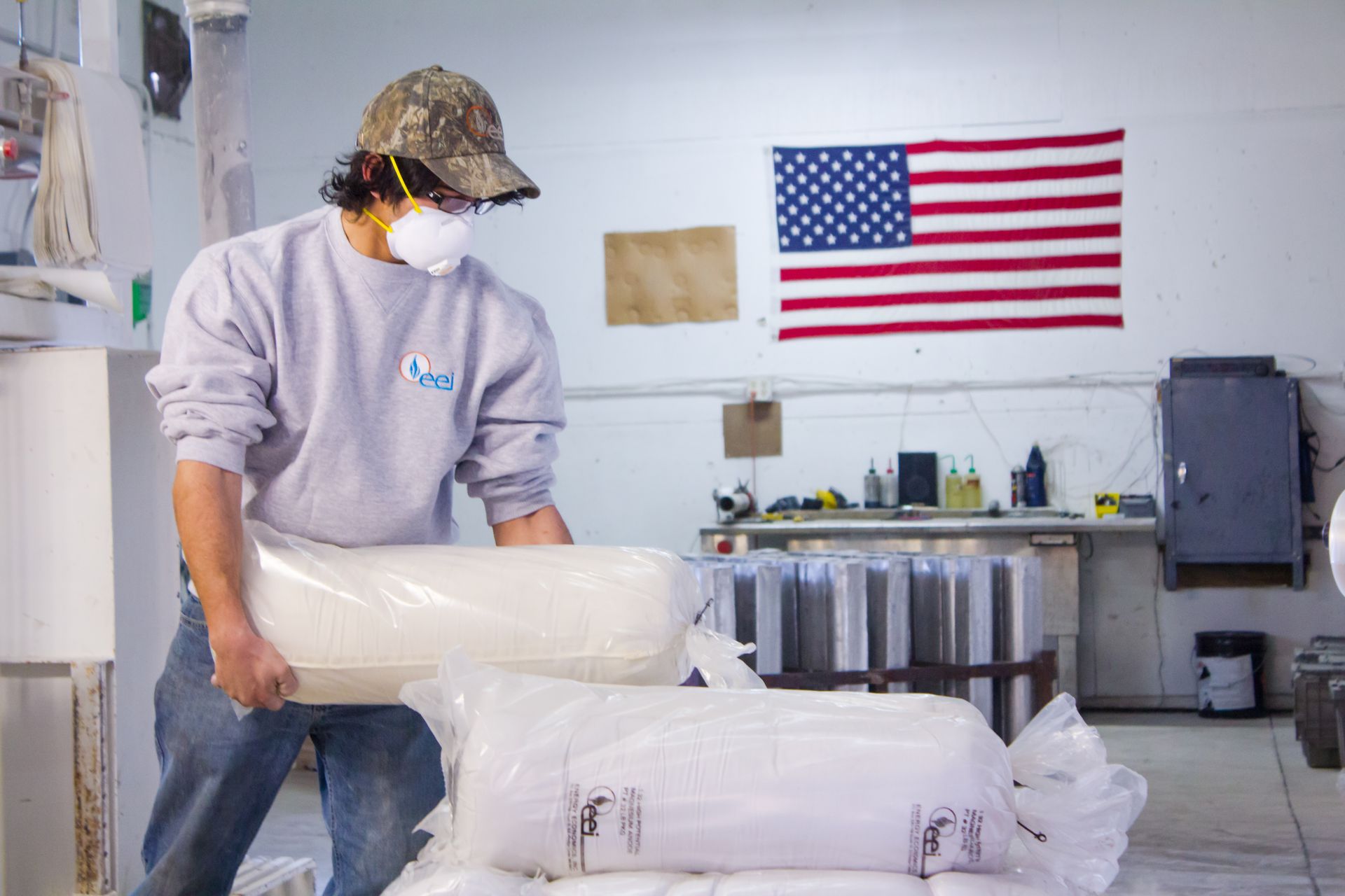 Person wearing a mask and hat lifting a bagged magnesium anode in a factory, with an American flag in the background.