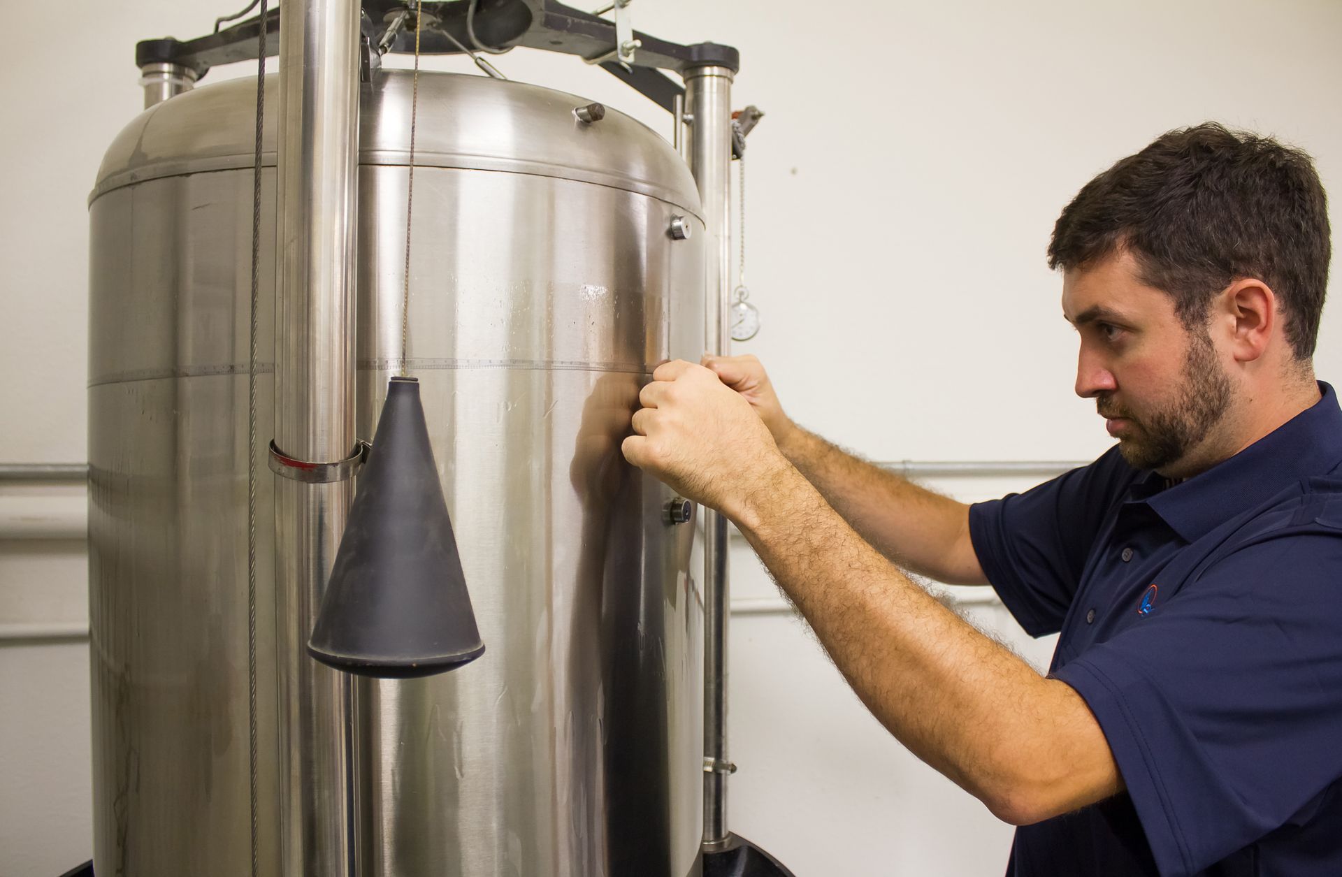 A man in a blue shirt works on a large stainless steel tank in a laboratory. He is calibrating a bell