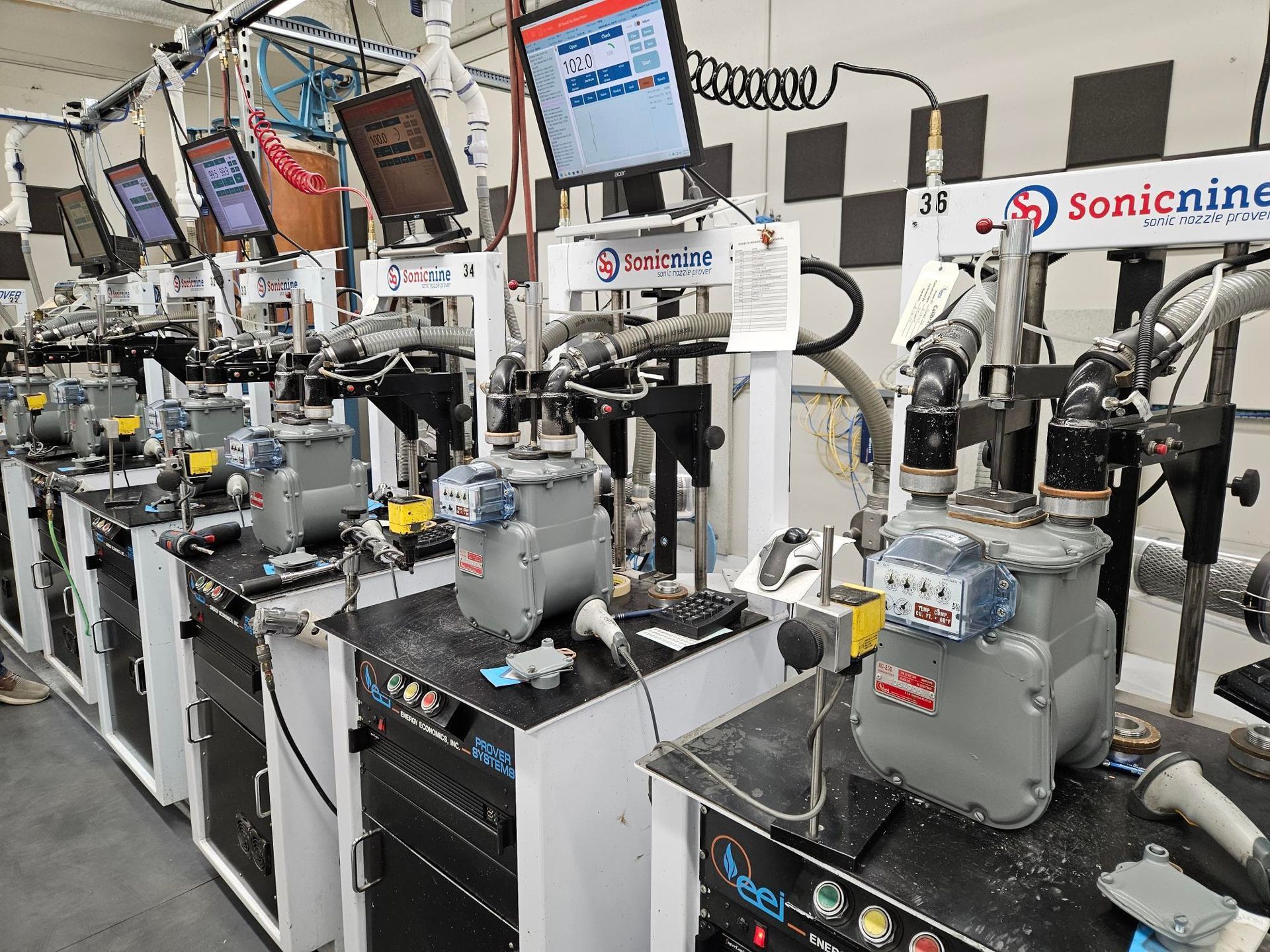 Assembly line with several machines testing gray utility meters, each station with a computer screen and tools.