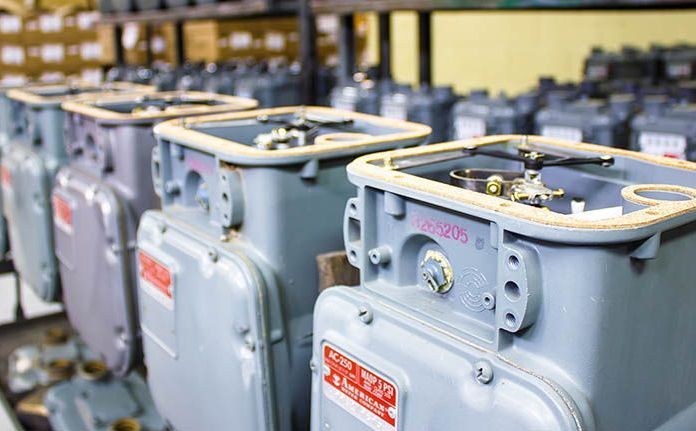 Rows of gray gas meters with open tops, in a storage or warehouse setting. Red and white labels are visible on some meters.