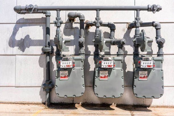 Three gray gas meters mounted on a wall with associated piping and valves, lit by sunlight.