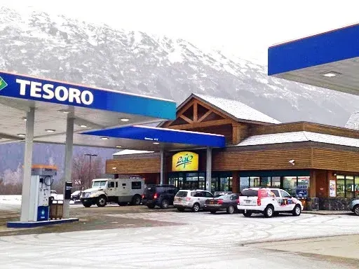A Tesoro gas station with wooden architecture set against a snowy mountain backdrop, with vehicles parked out front.