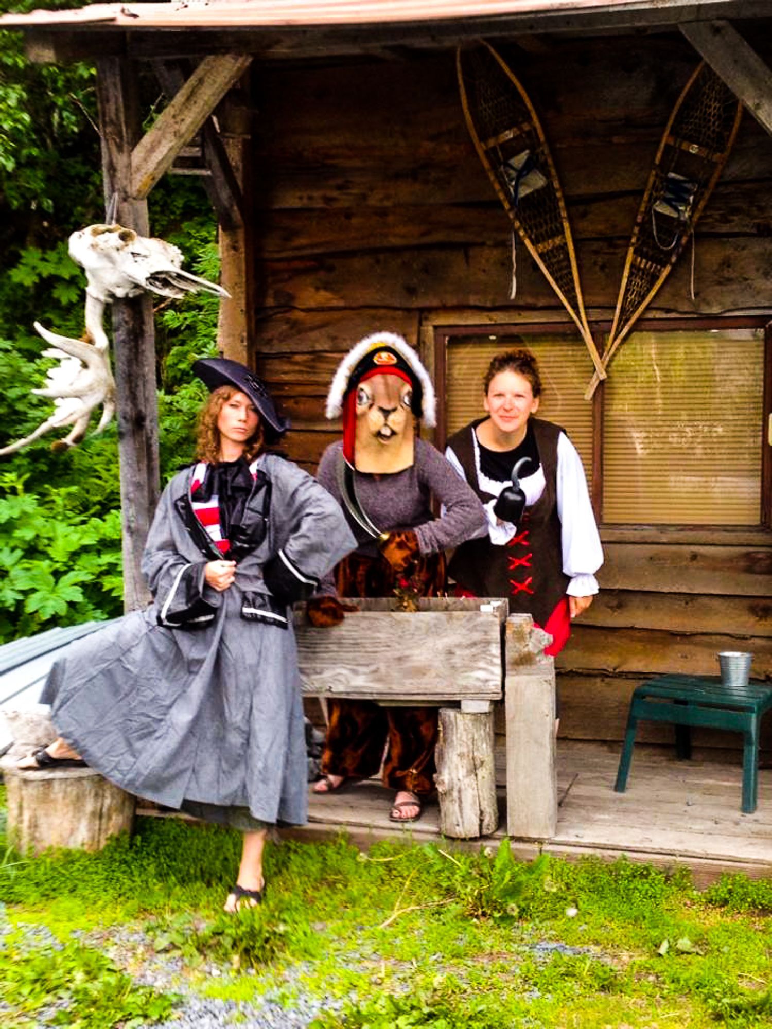 Three people in costumes pose on a porch. One wears a hat and long dress, the other a bear mask, and the third a vest.